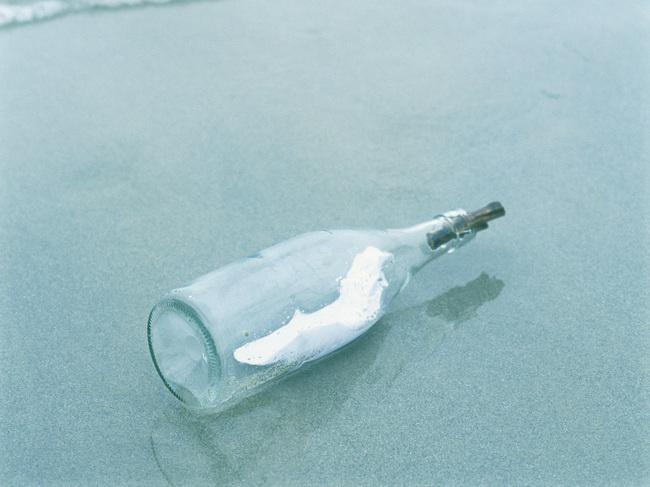 Eine Glasflasche liegt am Strand, umgeben von sanften Wellen und feinem Sand. In der Flasche ist ein dünnes Papier.